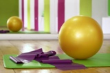 Auf dem Boden einer Sporthalle liegen ein Gymnastikball, eine Matte und ein Fitnessband.
