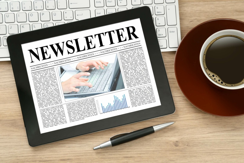 Auf einem Schreibtisch liegt eine Computertastatur, ein Tablet mit dem Schriftzug "Newsletter", sowie ein Kugelschreiber und eine Tasse Kaffee.