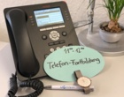 Auf einem Schreibtisch steht ein Telefonapparat mit einer Zettelnotiz "11:00 - 12:00 Telefonfortbildung". Daneben liegen eine Armbanduhr und ein Kugelschreiber.