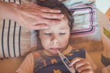 Ein Kind liegt im Bett mit einem Fieberthermometer im Mund und eine Hand befühlt die Stirn.
