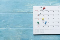 An einer blauen Holzwand hängt ein Monatskalender mit einigen Pinsteckern.