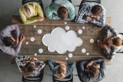 Von oben betrachtet, sitzen acht Personen um einen Tisch herum und schauen auf die Tischplatte, auf der eine Gedankenwolke ist, Symbolbild für Teammeeting und Ideenfindung.