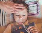 Ein Kind liegt im Bett mit einem Fieberthermometer im Mund und eine Hand befühlt die Stirn.