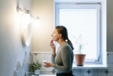 Eine Frau steht im Badezimmer vor einem Spiegel und cremt sich das Gesicht ein.