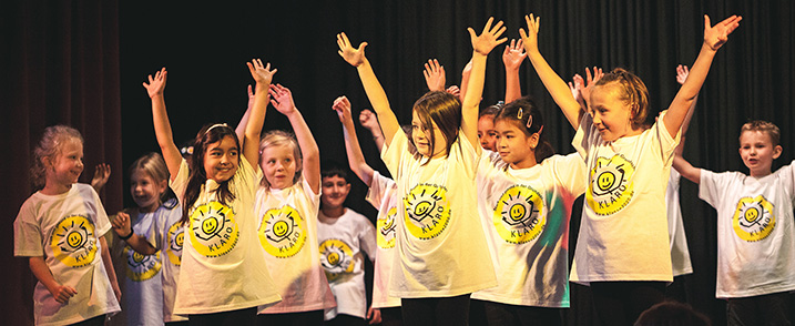 Kinder stehen auf einer bühne und recken die Arme hoch, Alle tragen das gleiche T-Shirt.