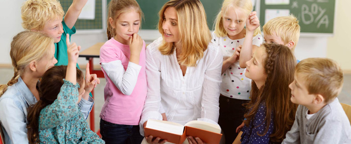 Eine Lehrerin ist umringt von Grundschulkindern in einem Klassenraum und liest aus einem Buch vor.